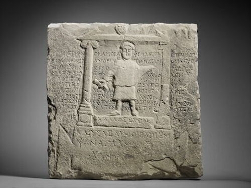 photo of the ancient Greek marble bas-relief tombstone of Aphrodisios, showing the subject standing between two pillars holding two items (a scroll, probably, and something else), with text behind and under him