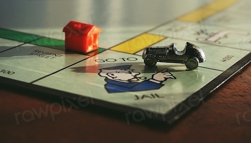 Close-up photo showing the edge of a monopoly board with the race car playing piece on the Go To Jail square and a small red plastic house on an adjacent square 
