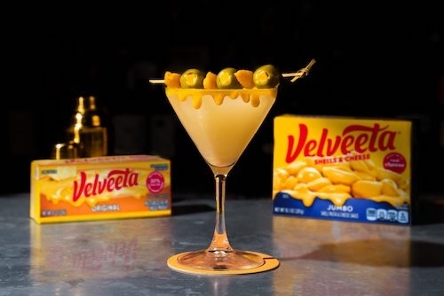 photo of of orange liquid in martini glass with a spear of olives and cheese and two boxes of velveeta in the background