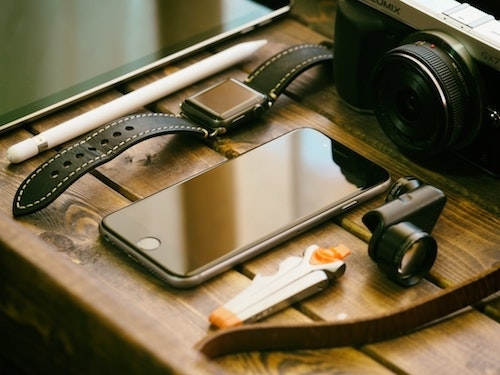 Close-up photo of camera, tablet, tablet pen, watch, mobile phone, multi-tool, loupe (?), and leather strap all arrayed in a pleasing composition on a nice wooden table top