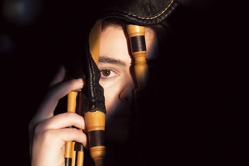 Somber close-up of Brighde Chaimbeul peering out through the pipes of a bagpipe or smallpipe instrument