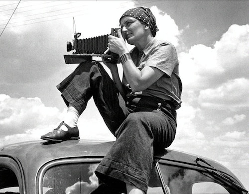 Dorothea Lange black and white self portrait showing the photographer casually dressed in trousers and kerchief perched atop a car shooting a photo with a large format camera