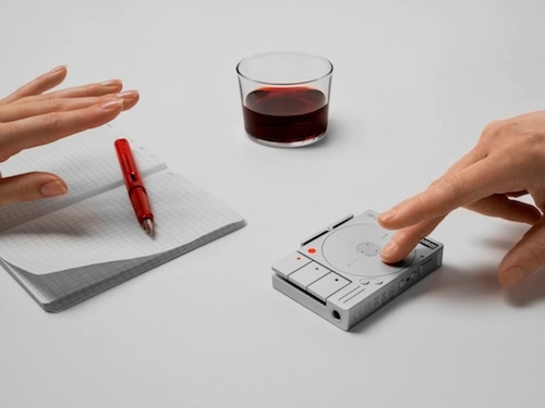 Photo of a pair of hands with a small, open, field notebook and fountain pen on the left and small, sleek, chrome colored TP–7 audio recorder on the right with a glass of red wine in the middle back