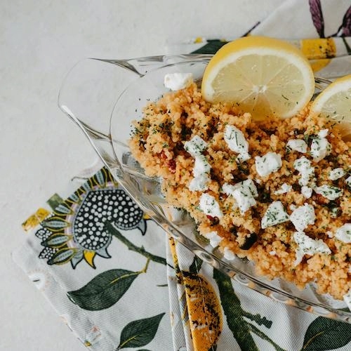 a glass serving bowl of couscous dotted with dollops of white cheese and half a lemon sitting atop a colorful napkin on a table