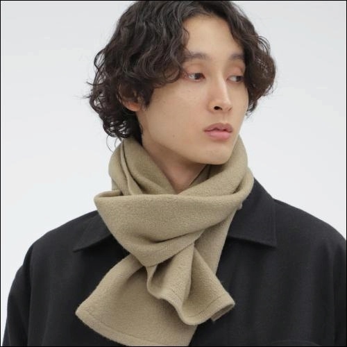 Fashion image of a model with short, wavy, dark hair wearing a beige fleece scarf tied or arranged attractively around their neck. They are dressed in a black coat and are looking off to the side against a plain white background.