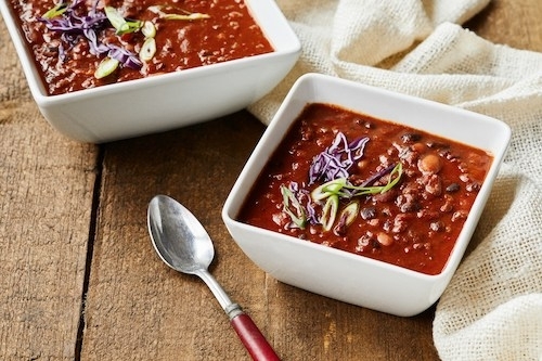 two bowls of chili in white, square bowls sitting on a wooden tabletop along with a cloth napkin and a spoon