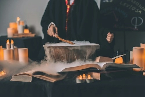 a person in a black robe surrounded by books and candles holding a wand over a cauldron emitting a foggy vapor