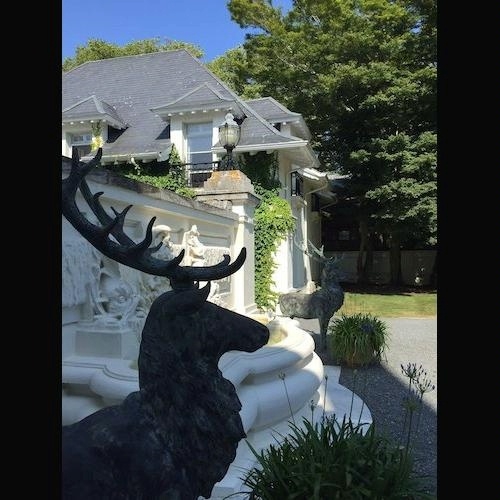 In the foreground, the visible head of a deer statue with a white, ivied mansion and dark wooded area in the background 