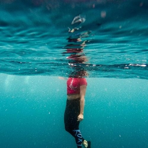 underwater photo of a woman in athletic-style red cropped top and black leggings treading water, head above the water's surface and body below