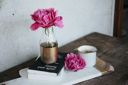 on a rustic wooden table, a cup of coffee, a widemouth glass bottle with a bright pink peony, and a stack of two books, the top one titled Milk and Honey