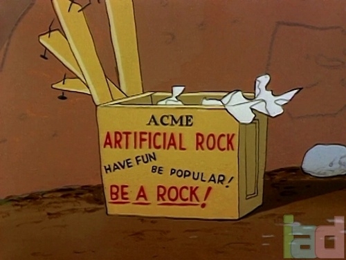 screenshot from a Roadrunner cartoon showing an unpacked wooden crate labeled ACME ARTIFICIAL ROCK / HAVE FUN / BE POPULAR! / BE A ROCK!