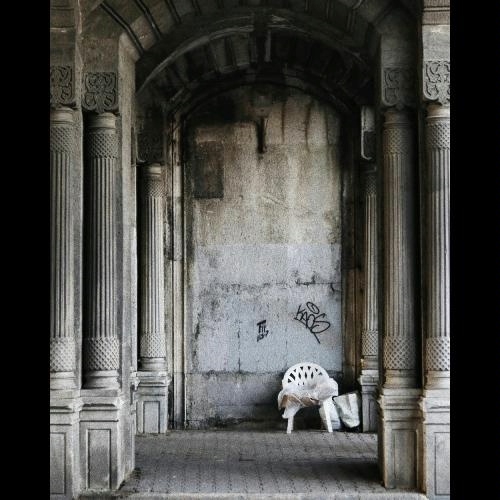 Black and white outdoor photo of beautiful, carved, weathered ancient columns converging toward a tall, rough-surfaced, stained, cracked, and graffitied gray wall with a white plastic lawn chair and plastic bags at the bottom