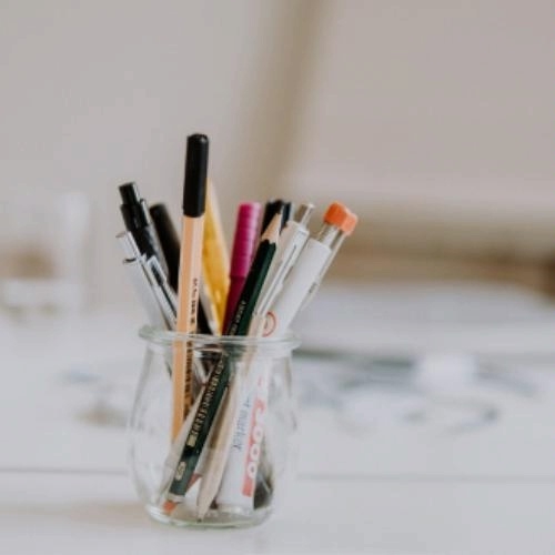 photo of a small glass jar of pens, pencils, and other drawing utensils, with a blurred background of a work in progress