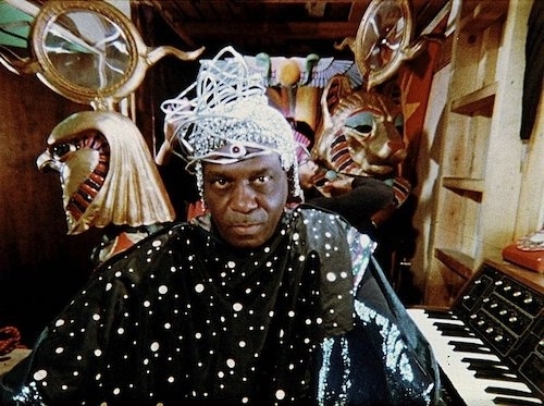 Photo of Sun Ra, dressed in a celestial-themed outfit adorned with stars and wearing an intricate metallic headpiece, sits at a keyboard surrounded by Egyptian-inspired sculptures of deities and mystical artifacts in a vibrant, otherworldly setting