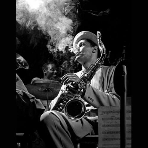 Black and white photo of Dexter Gordon sitting on stage as he exhales a plume of smoke during a break, 1948. He is wearing a stylish light colored suit, white shirt and pork pie hat. His saxophone is on his knee, and his left hand, holding a cigarette, rests against the instrument.