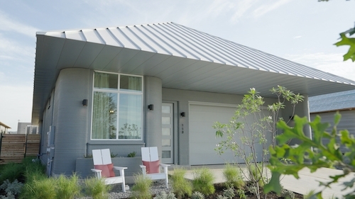 photo of a modern, single-story house with a sleek, slanted metal roof. The house features large windows, a simple front door, and smooth, curved walls. Two white chairs with red cushions are placed on the front porch, and the surrounding landscape includes various green plants and shrubs. The overall design is minimalist and contemporary, with clean lines and neutral tones.