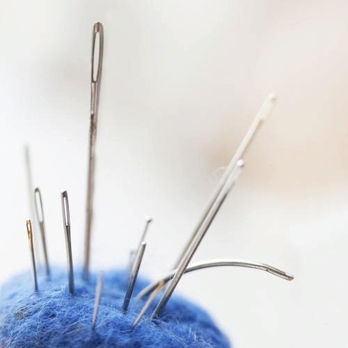 Close-up image of sewing needles of various sizes inserted into a blue pincushion. The needles are arranged at different angles, with a soft, blurred background emphasizing the sharpness of the needles and the texture of the pincushion.