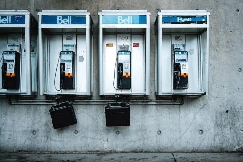 A row of four Bell-branded phone booths mounted on a concrete wall, each containing a payphone with black handsets and coin slots. The two booths on the left have black metal cases hanging below them. The image features a slightly worn, urban aesthetic with visible scratches and marks on the booths and wall, emphasizing a sense of decay or obsolescence.