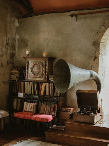 The image shows a cozy, vintage interior space within the Manor at Hemingford Grey. It features an antique phonograph with a large, ornate brass horn placed on a wooden platform. Surrounding the phonograph are shelves filled with books, a patterned cabinet, and two upholstered chairs with vibrant red and striped cushions. The walls are textured and aged, adding to the historic charm, and a warm light filters in from a nearby arched window.