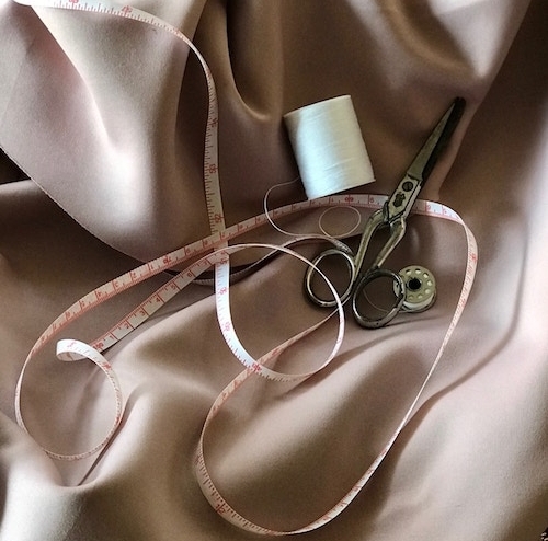 on top of taupe colored fabric, a white and red measuring tape, white spool of thread, bobbin with white thread, and a pair of scissors