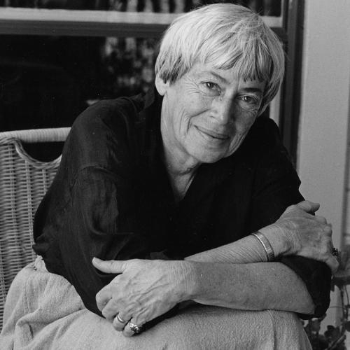 black and white photo of Le Guin in dark blouse and light colored skirt sitting in front of a bookcase, arms on knees, smiling at camera