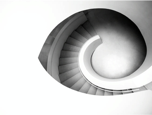 A black-and-white photograph of a spiral staircase taken from above, showing its elegant curvature. The image has high contrast, with the steps appearing dark and the outer area stark white. The design creates a smooth, circular pattern resembling an abstract eye.