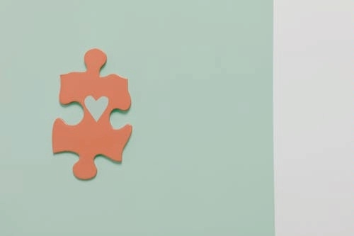 a single salmon-colored jigsaw puzzle piece with a pale green heart printed on it, against a pale green background