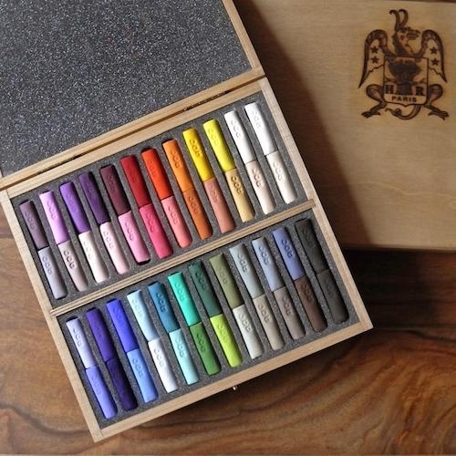 At an angle, a wooden box of 48 pastel half-sticks in two trays of four rows, with top ranging from puce to red to yellow to white, and the bottom tray ranging from lilac to teal to green to grey to black. The sticks are noticeably handmade and each stamped ROC. Part of the box's wooden lid can be seen to the right, with fanciful dragon crest and PARIS emblazoned on it.