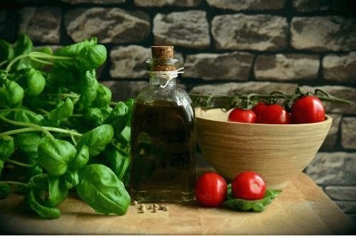 The image shows a rustic food setup featuring a bundle of fresh basil leaves on the left, a small glass bottle filled with olive oil and sealed with a cork in the center, and a wooden bowl containing vine-ripened cherry tomatoes on the right. There are a few loose tomatoes, some placed on basil leaves, in front of the bowl. The background is a stone wall, giving the scene a natural, earthy feel, with a focus on fresh ingredients commonly used in Mediterranean cooking. The lighting is soft and warm, adding a cozy ambiance.