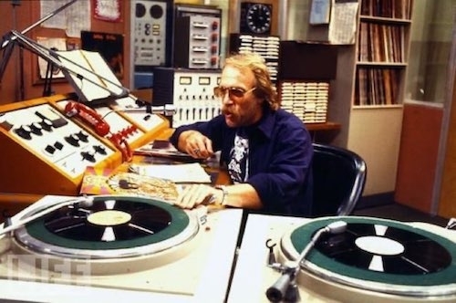 Johnny Fever behind his DJ desk with turntables in the foreground and audio equipment, tapes and LPs in the background