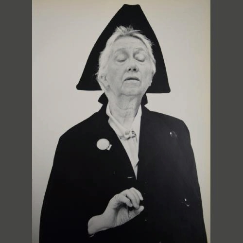 Black and white photo of an elderly woman wearing a dark coat and a distinctive triangular hat. Her eyes are closed, and one hand is raised with a poised gesture, conveying a serene or reflective expression