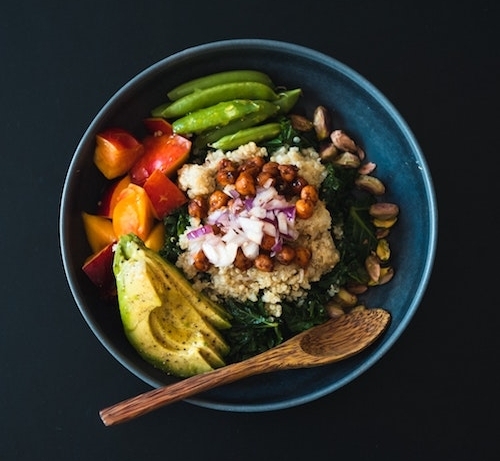 A blue bowl with a small wooden spoon on a black background. In the bowl are snow peas, sliced avocado, tomatoes, cooked greens, quinoa, and sauced chickpeas