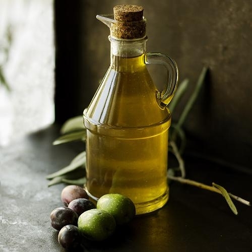 a clear glass cruet bottle of olive oil on a stone surface with a few green and black olives in front and an olive branch behind