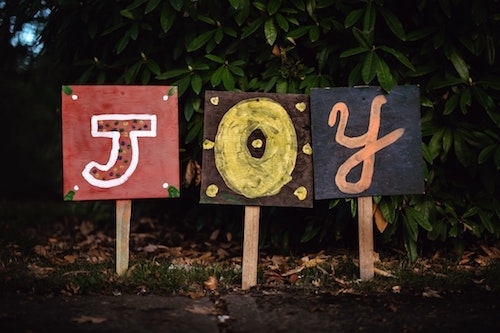 Three colorful homemade signs on wooden stakes driven into the ground, on each sign a different letter J-O-Y, each in a different style