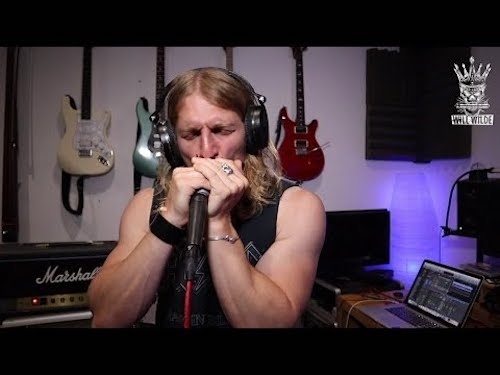 A man with shoulder-length blonde hair and black sleeveless t-shirt in studio setting playing harmonica with intense concentration