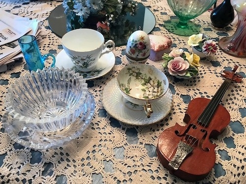 a collection of items on a polished wood tabletop covered by a crocheted cloth, including tea cups and saucers, a violin, an egg cup, a cut glass bowl, china candleholders, a tray, and old newspapers