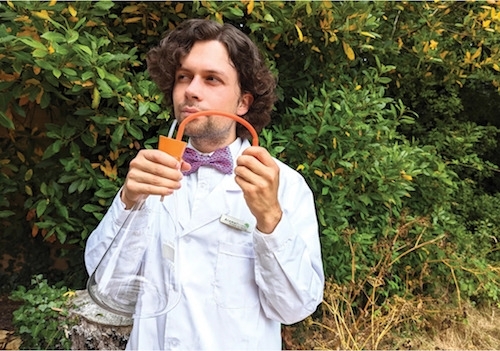photo of Heritage Scenting Consultant Liam R Findlay in front of a green, leafy background, wearing a white lab coat and polka dot bow tie, and holding a glass lab flask topped by what may be a bulb atomizer device. He wears a thoughtful, satisfied expression while seeming to sniff from the atomizer.