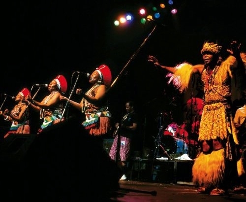 photo: performing onstage, male performer Mahlathini in tribal garb and three female musicians, dressed in original attire as young Zulu girls – but with red izicholo on their heads to signify they were now grown women: Nobesuthu Shawe, Hilda Tloubatla and Mildred Mangxola