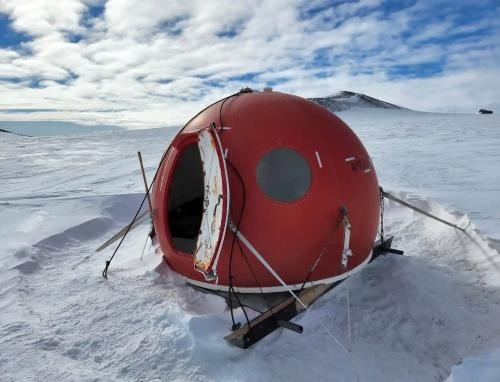 Daylight photo of a round red apple-like pod structure in foreground with a background of a mostly flat, empty snowy expanse and beautiful fluffy clouds spreading out over a huge sky