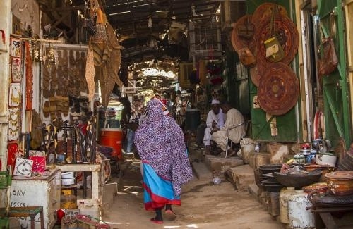 a narrow alley is lined with small shops and stalls displaying a variety of traditional goods, including woven baskets, leather sandals, pottery, and decorative items. A woman wearing a colorful patterned shawl and a bright red dress walks through the market, adding movement and liveliness to the scene. Several men, dressed in white traditional attire, sit on steps in the background, engaging in conversation. The atmosphere is bustling yet relaxed, with an array of textures, colors, and cultural artifacts