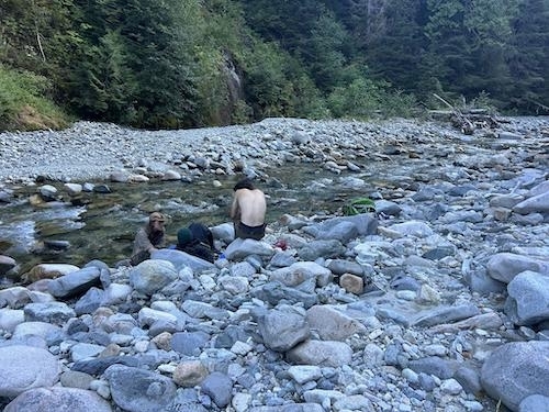 photo of two visible people on a rocky riverbed surrounded by large, smooth stones with a forested area in the background