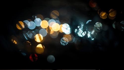 against a black background, an abstract, blurred collection of circular light spots, creating a bokeh effect. The lights are in various colors, including warm yellows, cool blues, and a few reds