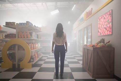 A woman in white shirt and trousers stands with her back to us facing an eerie, ominous, foggy supermarket setting