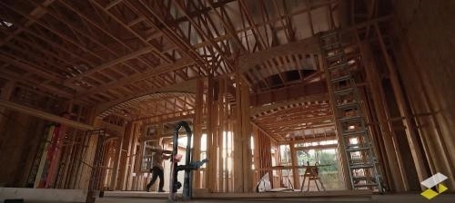 image showong the interior framing of a building under construction. The structure consists of exposed wooden beams, studs, and joists forming the walls and ceiling. Two curved archways are visible, emphasizing architectural detail. A person is standing near the center, possibly working with tools or inspecting the area. A ladder leans against one wall on the right side, and construction equipment is scattered throughout.