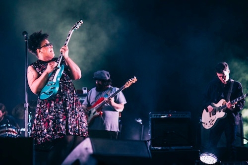 Night-shot photo of Brittany Howard onstage dressed in a sleeveless multicolor flowy dress and black eyeglasses, playing a bright blue-green Gibson electric guitar