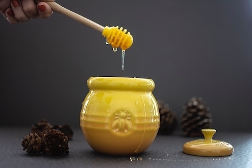 surrounded by several pinecones and lid, a vintage-style yellow ceramic honey jar with a hand holding a honey dripper above